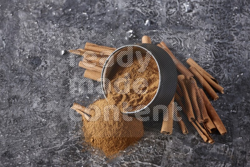 Black pottery bowl over filled with cinnamon powder and cinnamon sticks around the bowl on a textured black background