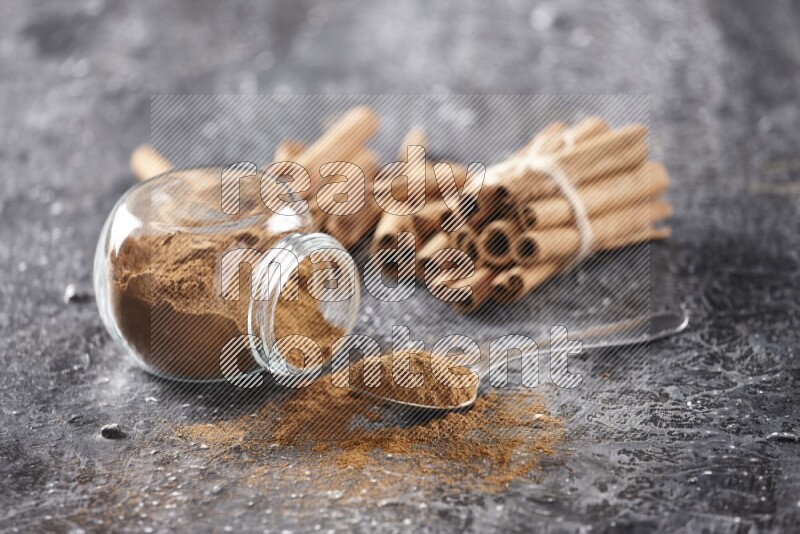 Herbal glass jar full cinnamon powder flipped and a metal spoon full of powder, cinnamon sticks stacked and bounded in the back on textured black background in different angles