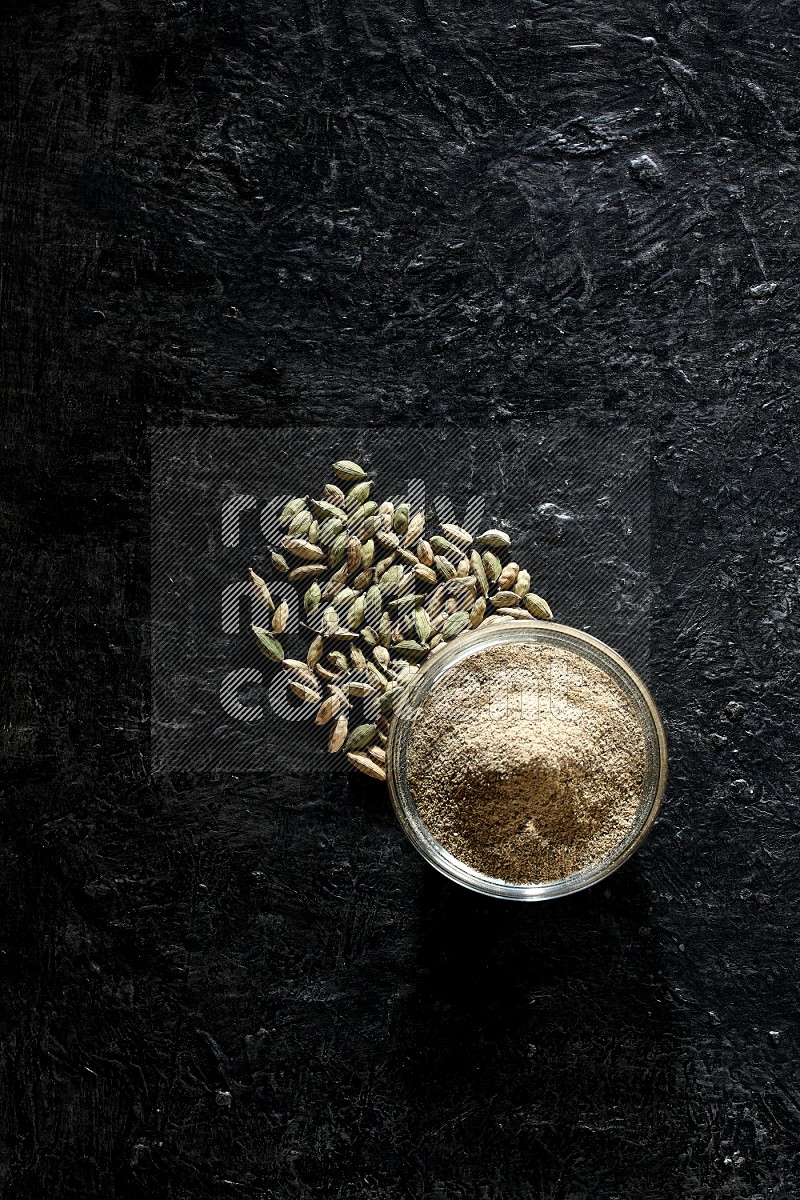 A glass bowl full of cardamom powder and cardamom seeds beside it on textured black flooring