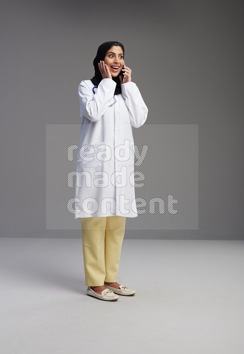 Saudi woman wearing lab coat with stethoscope standing talking on phone on Gray background