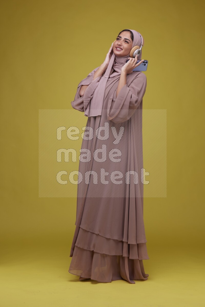 A woman Listening to Music on a Yellow Background wearing Brown Abaya with Hijab
