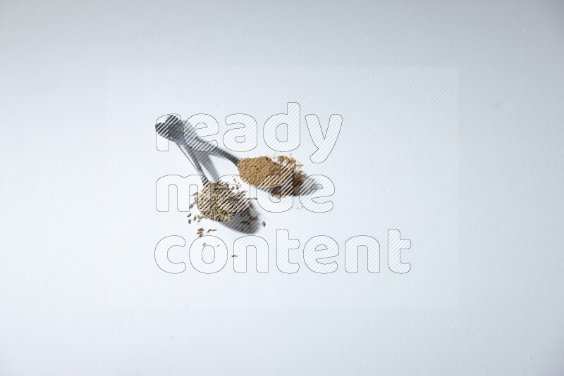 ملعقتان معدنيتان ممتلئتان بحبوب الكمون وبودرته على خلفية بيضاء