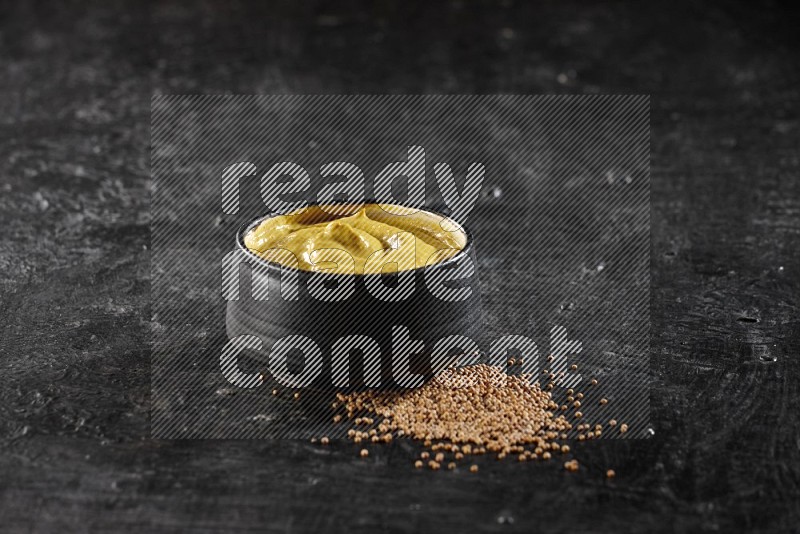 A black pottery bowl full of mustard paste with mustard seeds underneath on textured black flooring in different angles