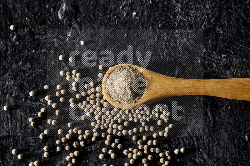 A wooden spoon full of white pepper powder with white pepper beads on textured black flooring