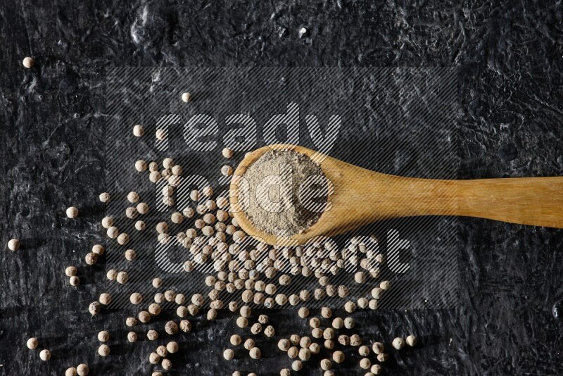 A wooden spoon full of white pepper powder with white pepper beads on textured black flooring