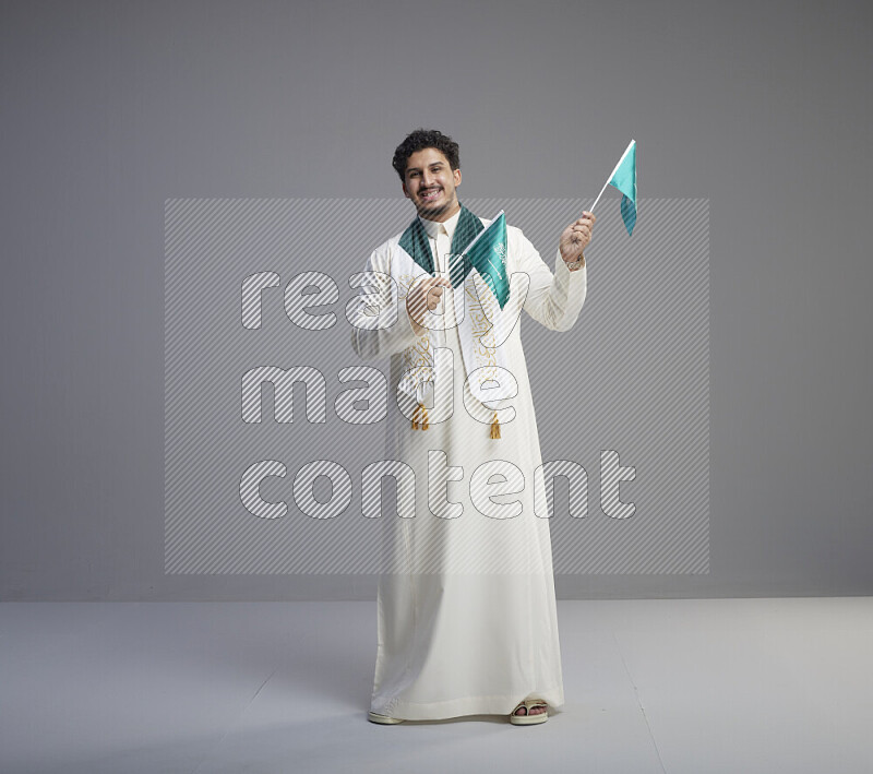 A Saudi man standing wearing thob and saudi flag scarf and holding small saudi flag on gray background