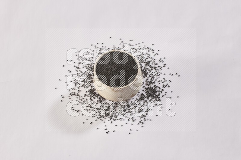 A beige pottery bowl full of black seeds and more seeds spread on a white flooring