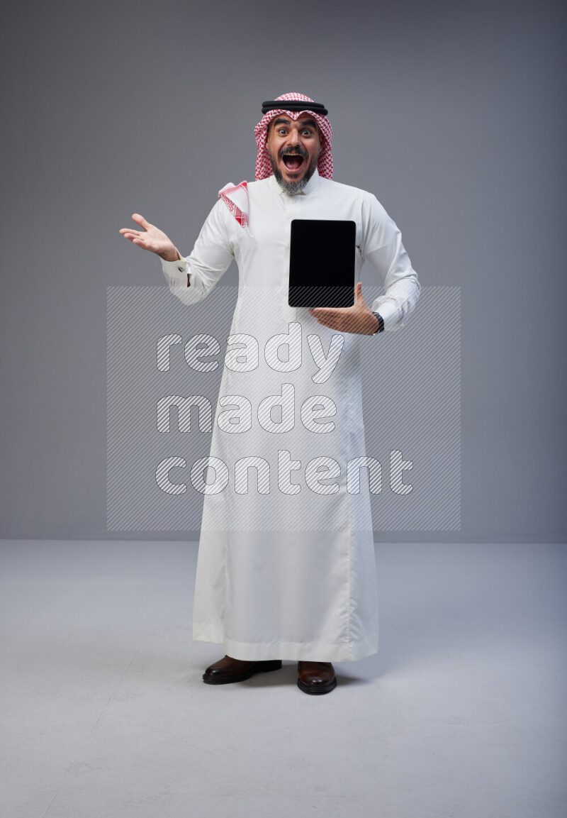 Saudi man Wearing Thob and red Shomag standing showing tablet to camera on Gray background