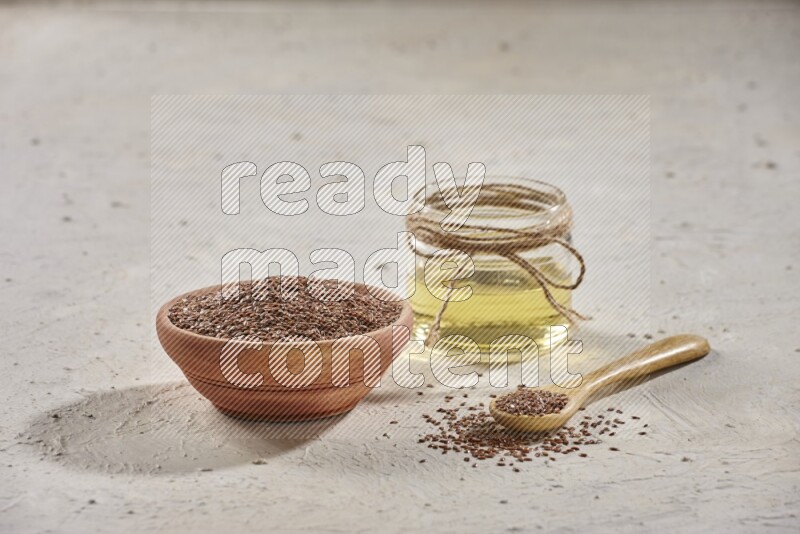 A wooden bowl and spoon full of flax seeds and a glass jar of flaxseeds oil on a textured white flooring
