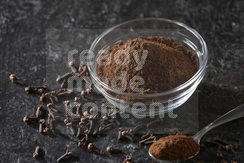 A glass bowl and a metal spoon full of cloves powder with gloves grains beside them on a textured black flooring