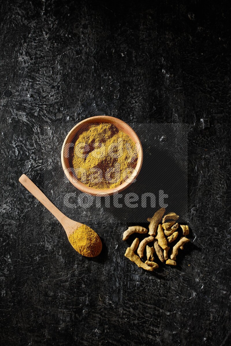 A wooden bowl and a wooden spoon full of turmeric powder with dried turmeric fingers on textured black flooring