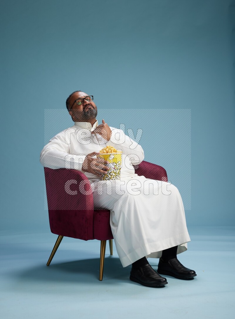 Saudi Man without shimag sitting on chair eating popcorn on blue background