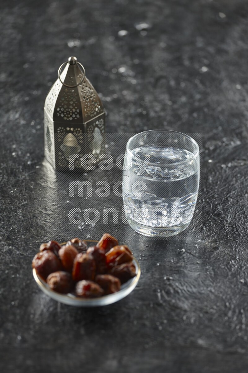 A silver lantern with different drinks, dates, nuts, prayer beads and quran on textured black background