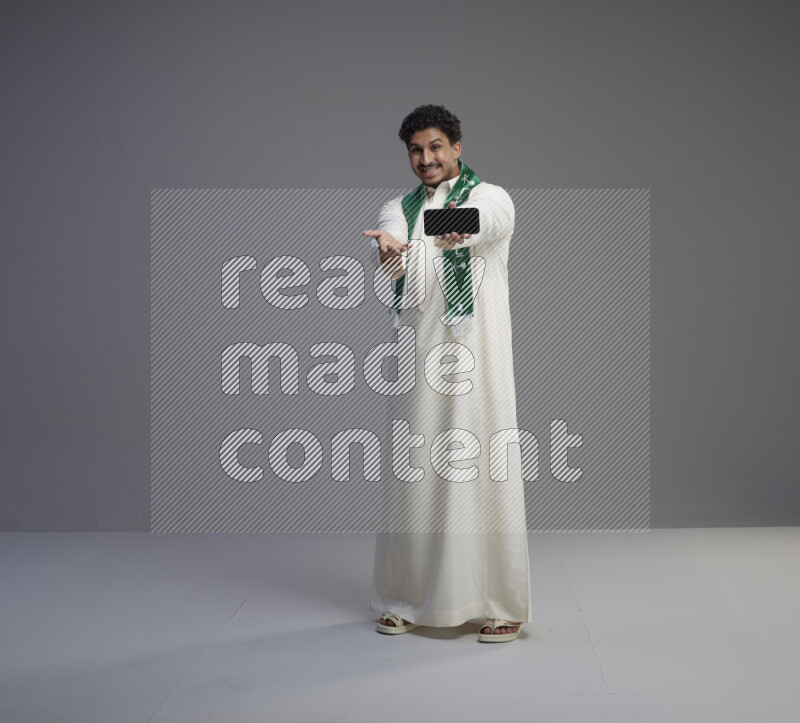 A Saudi man standing wearing thob and Saudi flag scarf showing phone to camera on gray background