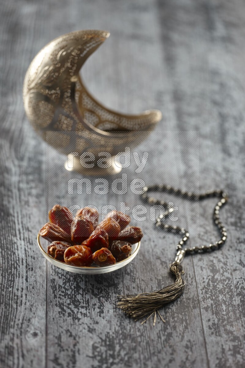 A silver lantern with different drinks, dates, nuts, prayer beads and quran on grey wooden background