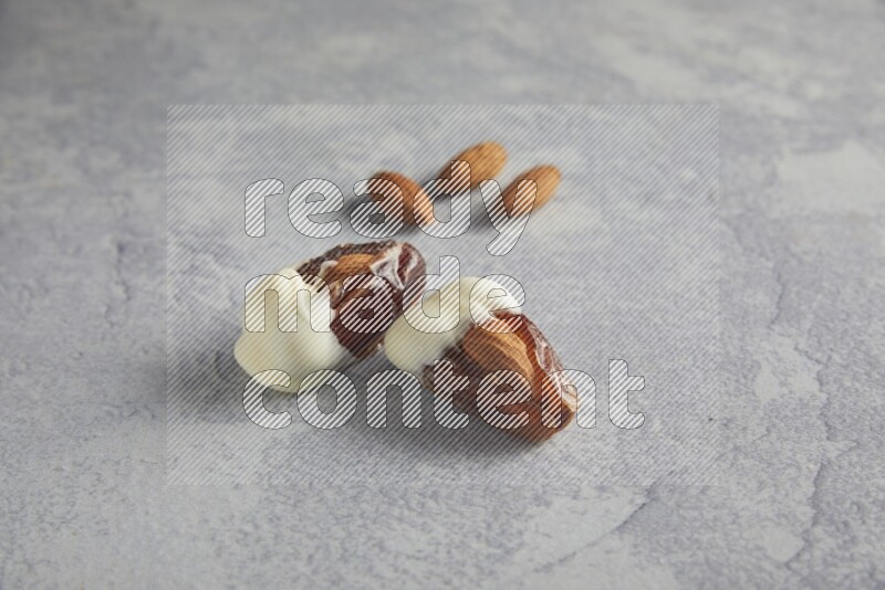 two almonds stuffed dates covered with white chololate with unroasted almonds on a light grey background