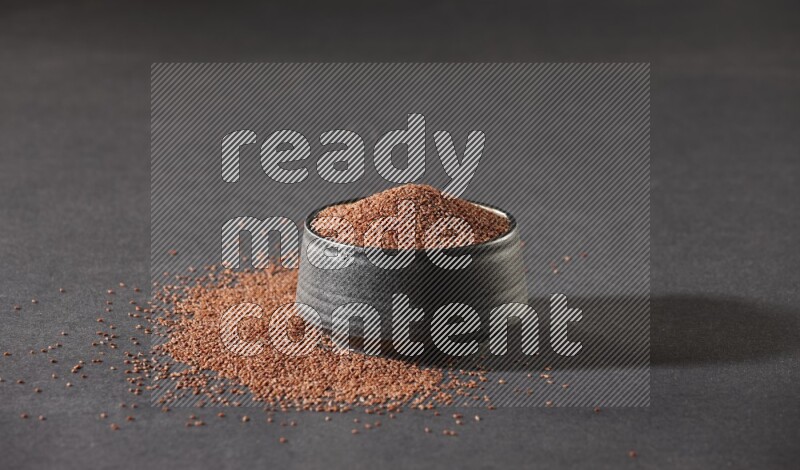 A black pottery bowl full of garden cress seeds surrounded by seeds on a black flooring