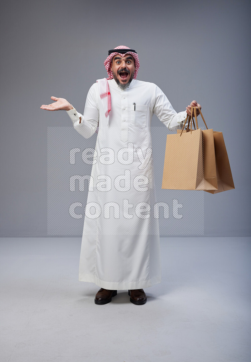 Saudi man Wearing Thob and red Shomag standing holding shopping bag on Gray background