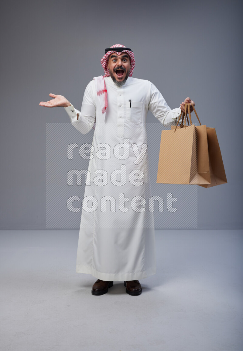 Saudi man Wearing Thob and red Shomag standing holding shopping bag on Gray background