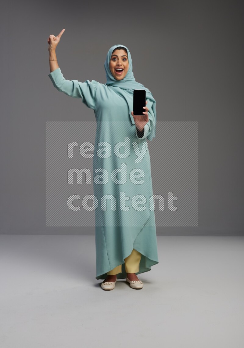 Saudi Woman wearing Abaya standing showing phone to camera on Gray background