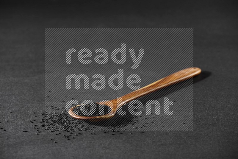 A wooden ladle full of black seeds and the seeds spread beside it on a black flooring