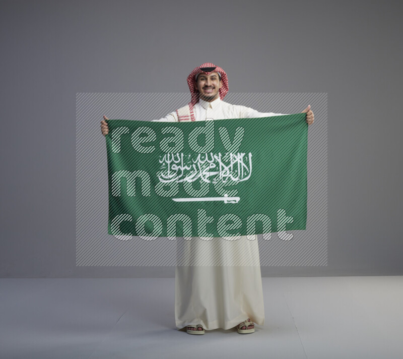 A saudi man standing wearing thob and red shomag holding big saudi flag on gray background