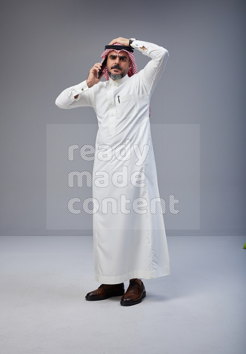 Saudi man Wearing Thob and red Shomag standing talking on phone on Gray background