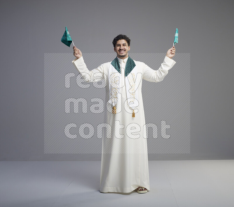 A Saudi man standing wearing thob and saudi flag scarf and holding small saudi flag on gray background