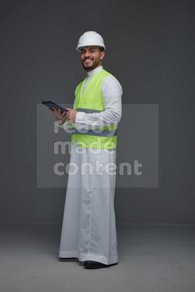A Saudi man wearing Thobe with a yellow safety vest and white helmet standing and using his tablet different angles eye level on a gray background