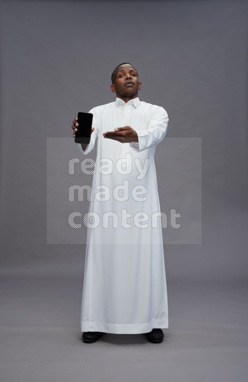 Saudi man wearing thob standing showing phone to camera on gray background