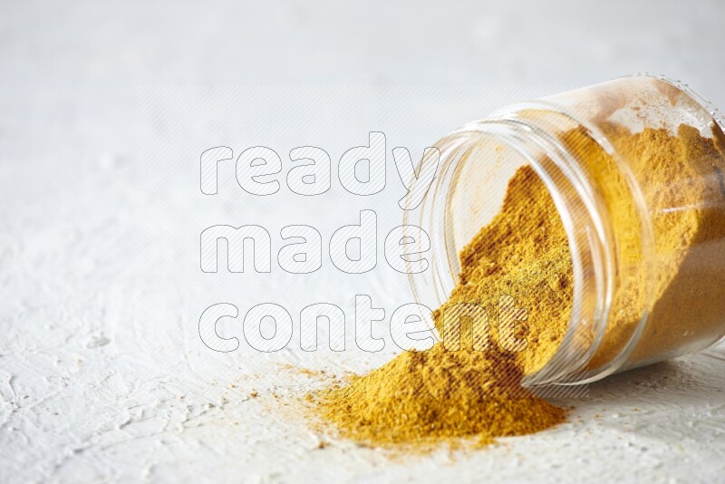 A flipped glass spice jar full of turmeric powder and powder fell out it on textured white flooring
