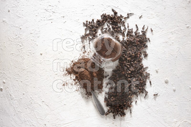 وعاء زجاجي مقلوب ممتلئ ببودرة القرنفل وتناثرت البودرة منه مع حبوب القرنفل المنتشرة على أرضية بيضاء