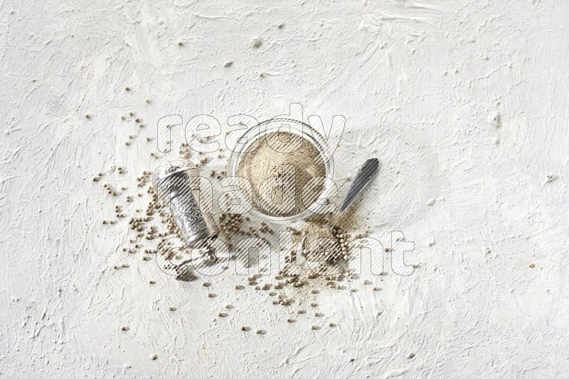A glass bowl full of white pepper powder with white pepper beads and a metal grinder on textured white flooring