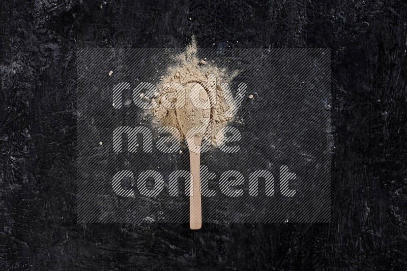 A wooden spoon full of garlic powder with scattered powder around it on a textured black flooring