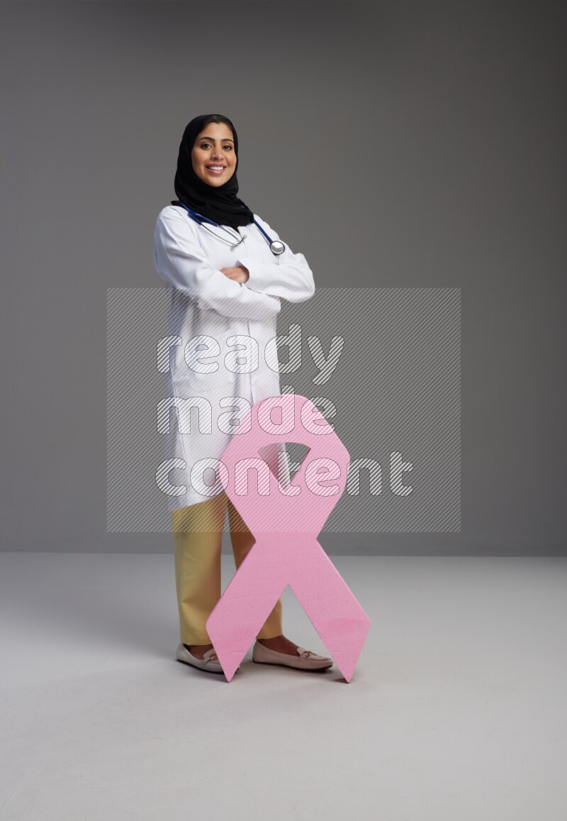 Saudi woman wearing lab coat with stethoscope standing holding awareness ribbon symbol standing on Gray background