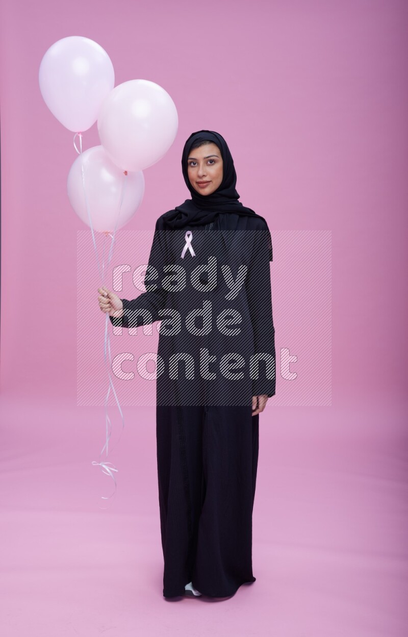 Saudi woman wearing pink ribbon on Abaya standing holding balloons on plain pink background