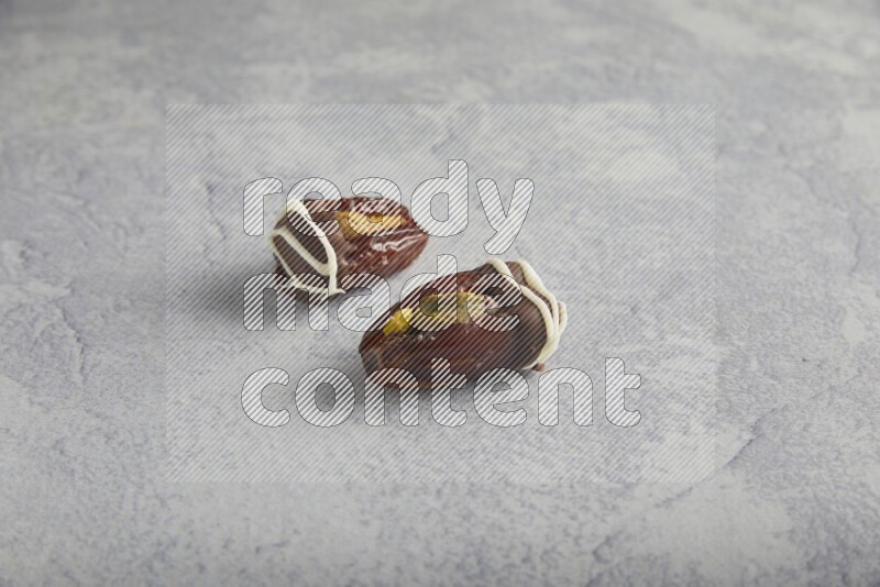 two pistachio stuffed dates covered with dark chocolate with white chocolate drizzle on a light grey background