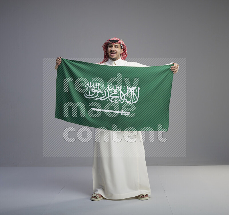 A saudi man standing wearing thob and red shomag holding big saudi flag on gray background