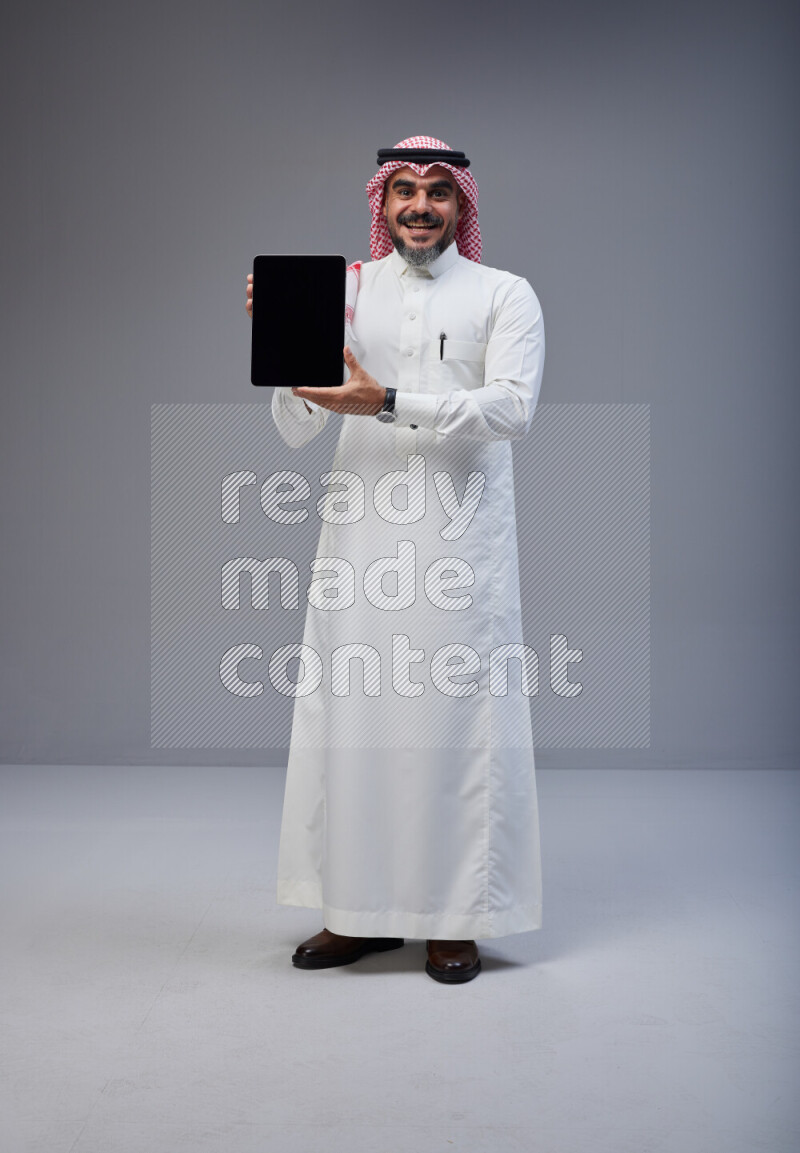 Saudi man Wearing Thob and red Shomag standing showing tablet to camera on Gray background