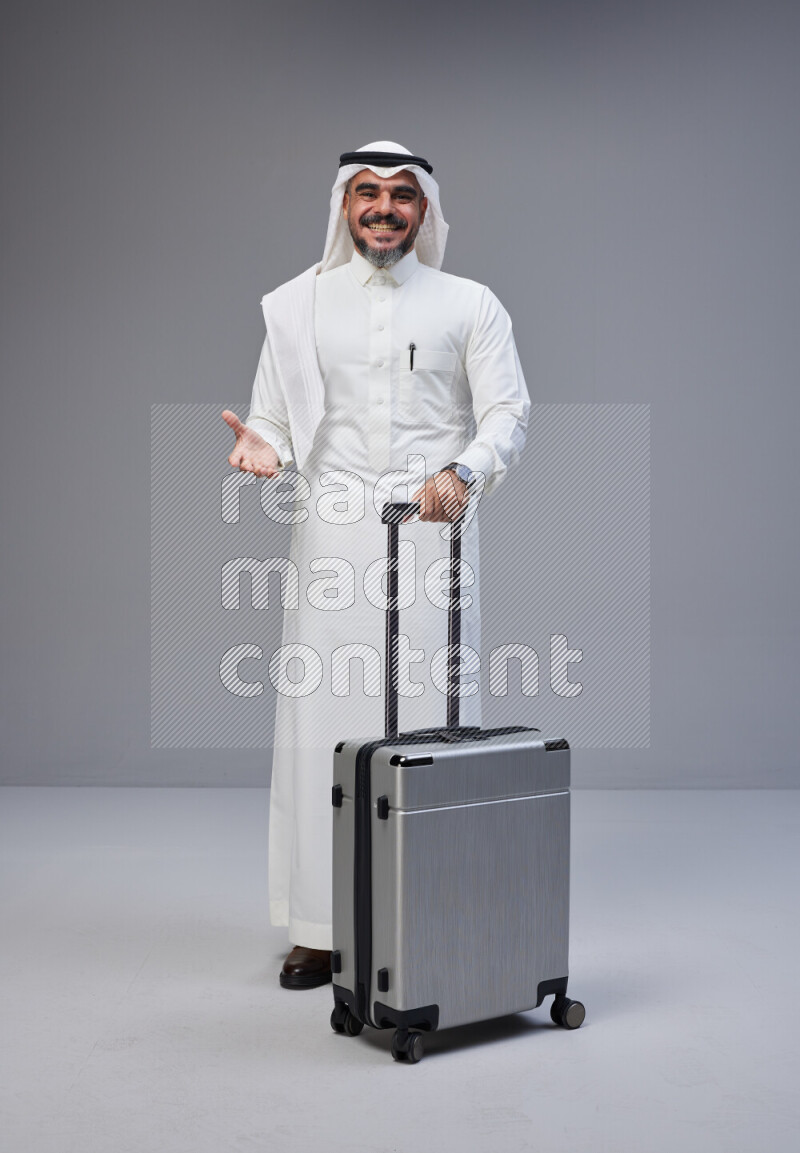 Saudi man wearing Thob and white Shomag standing holding Travel bag on Gray background