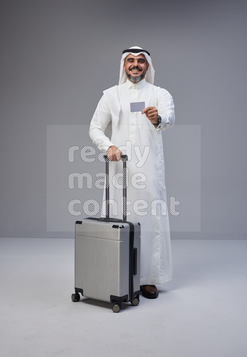 Saudi man wearing Thob and white Shomag standing holding Travel bag and ATM card on Gray background