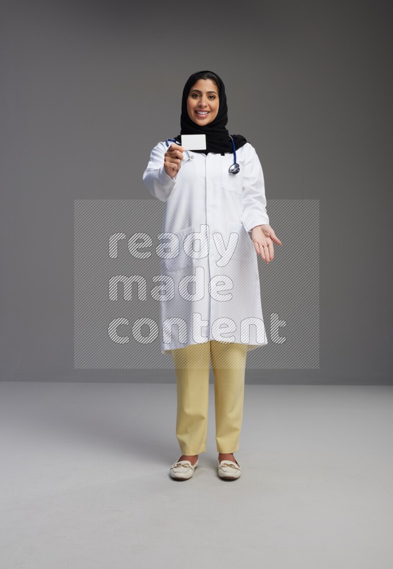 Saudi woman wearing lab coat with stethoscope standing holding ATM card on Gray background