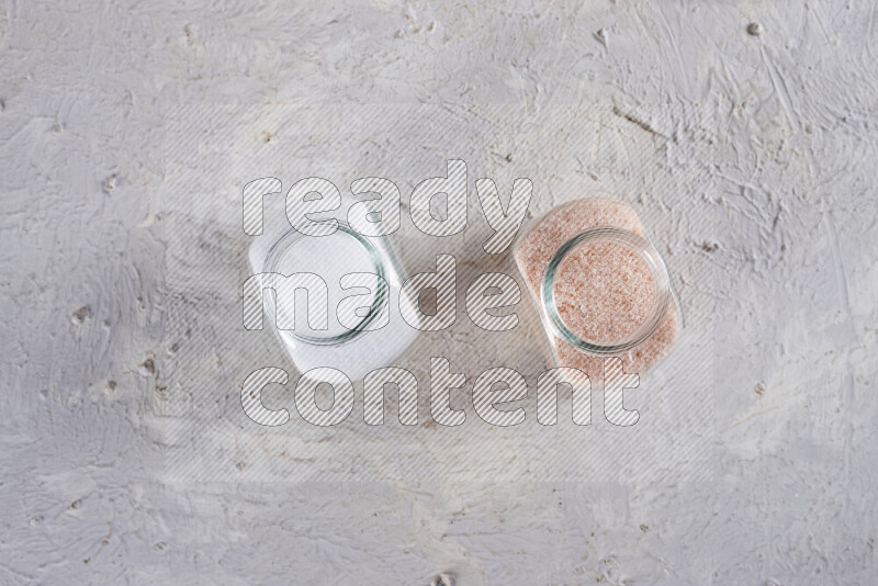 2 glass jars one is full of fine himalayan salt and the other with table salt on white background