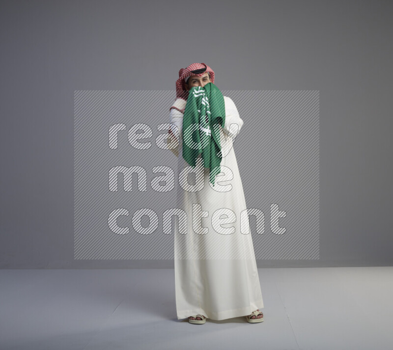 A Saudi man standing wearing thob and red shomag kissing big Saudi flag on gray background