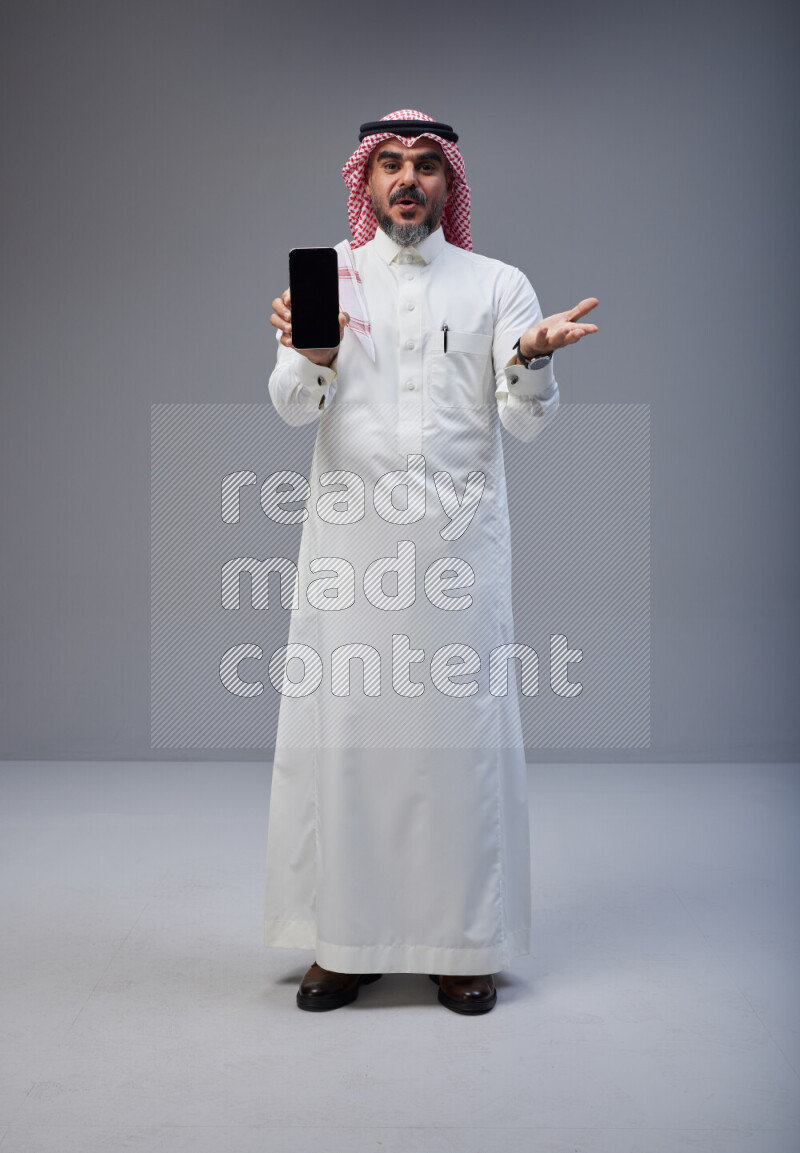 Saudi man Wearing Thob and red Shomag standing showing phone to camera on Gray background