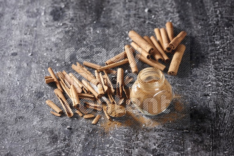 Herbal glass jar and a metal spoon full of cinnamon powder surrounded by cinnamon sticks on textured black background