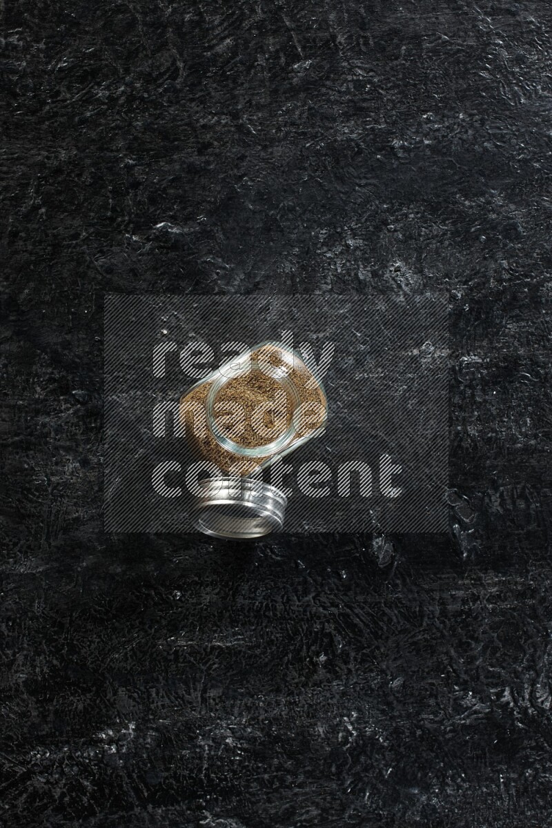 A glass spice jar full of cumin powder on a textured black flooring