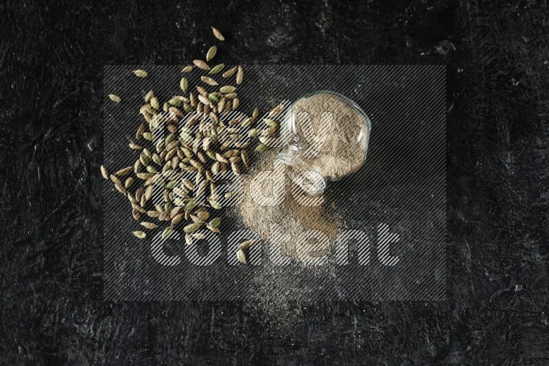 وعاء زجاجي للتوابل مقلوب وممتلئ ببودرة الهيل والبودرة تناثرت منه مع حبوب الهيل المتناثرة على خلفية سوداء