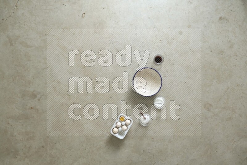 A ceramic bowl beside a carton of eggs and glass bowl full of sugar, vanilla extract, flour on beige marble background
