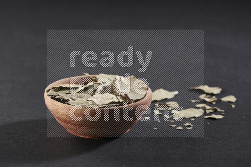 A wooden bowl filled with dried bay leaves on black flooring